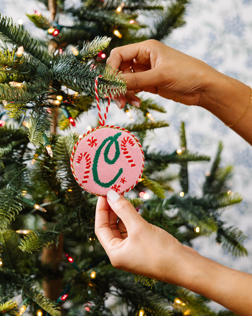 Hand holding a pink ornament with a green letter 'C' against a Christmas tree background