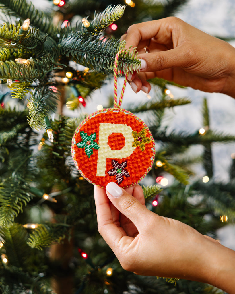 Hand holding a Christmas ornament with a letter 'P' in front of a decorated tree.