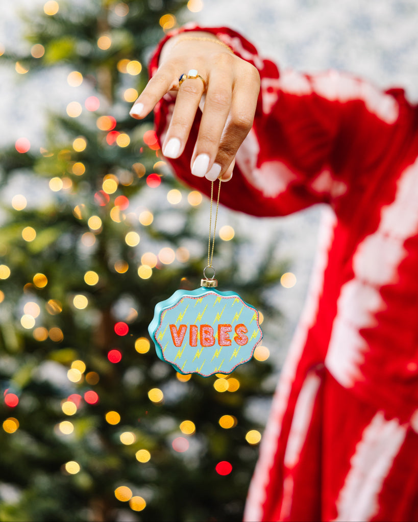 Hand holding a Christmas ornament with 'VIBES' text in front of a decorated tree.