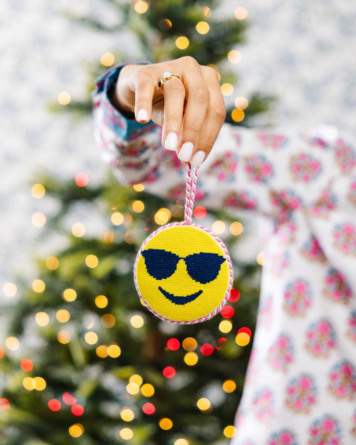 Hand holding a yellow emoji with sunglasses ornament in front of a decorated Christmas tree.
