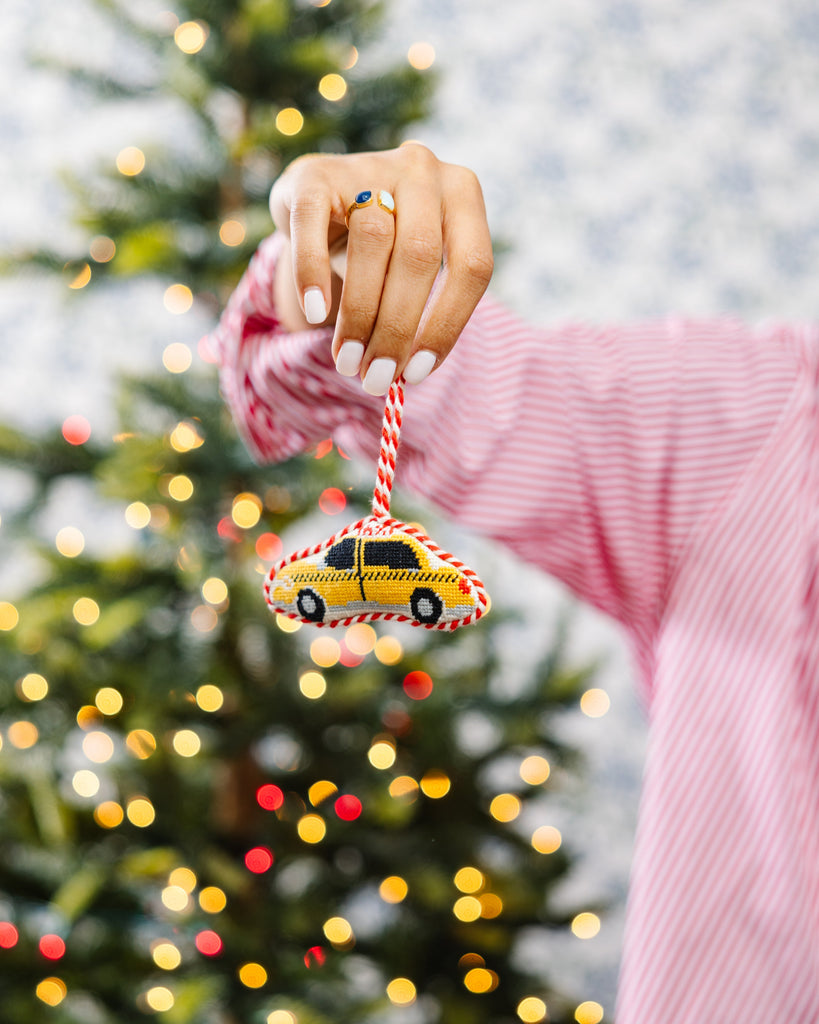 Person holding a Christmas ornament shaped like a yellow taxi cab in front of a decorated Christmas tree.