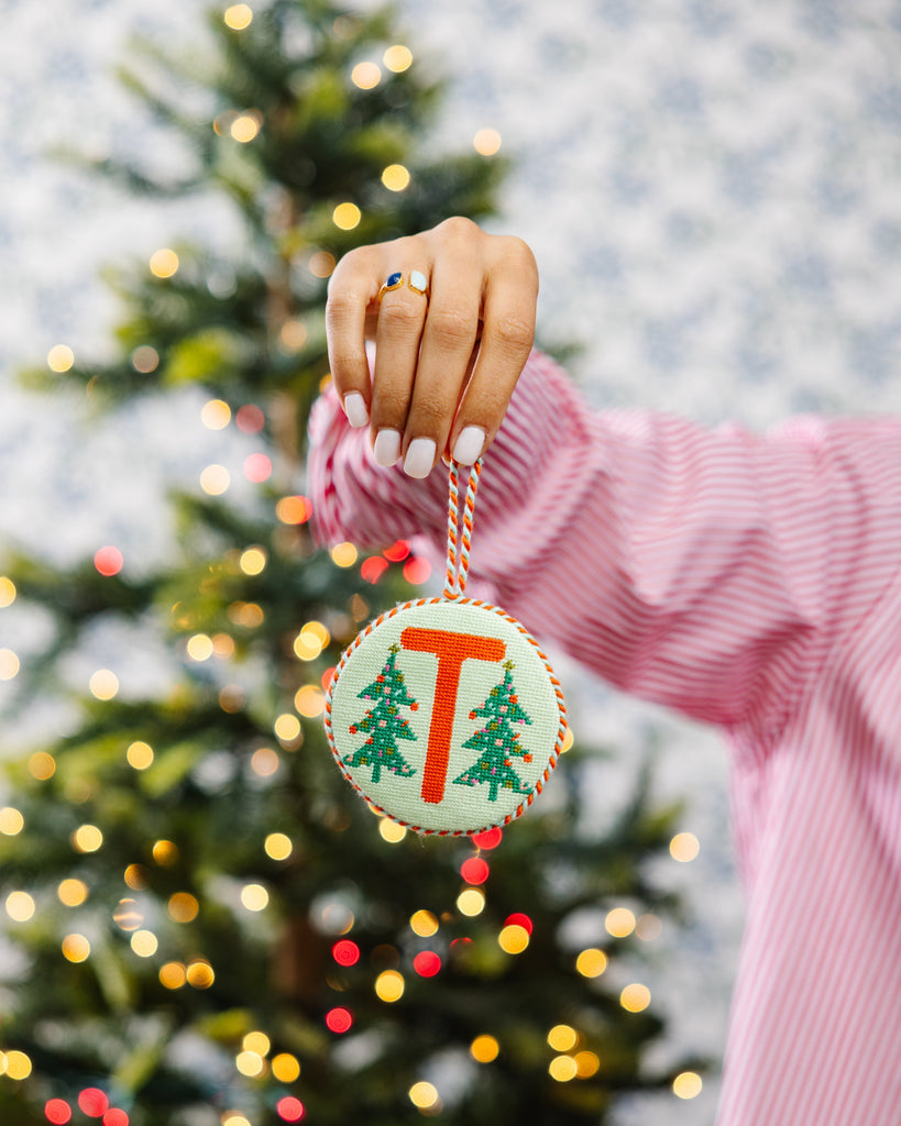 Hand holding a Christmas ornament with a tree design in front of a decorated Christmas tree.