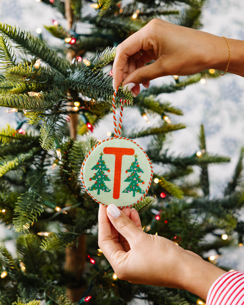 Hand holding a Christmas ornament with 'T' and tree design in front of a decorated tree.