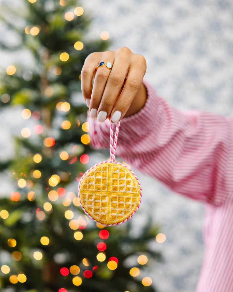 Hand holding a yellow waffle-shaped ornament with a blurred Christmas tree in the background