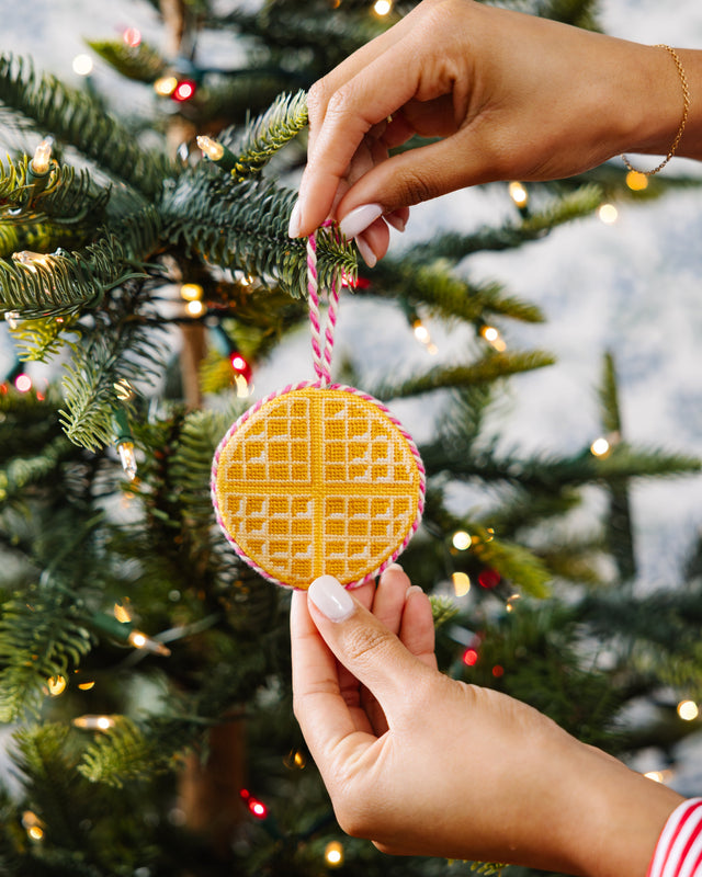 Hand holding a yellow cookie-shaped ornament on a Christmas tree with lights in the background