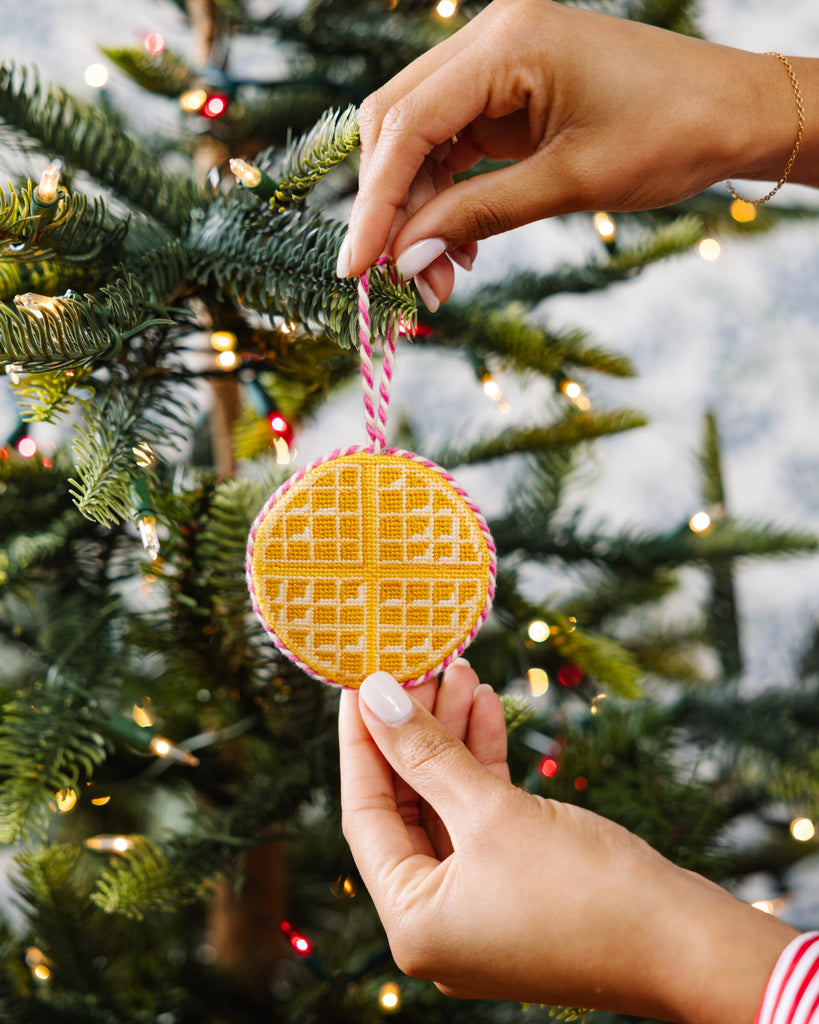 Hand holding a yellow cookie-shaped ornament on a Christmas tree with lights in the background