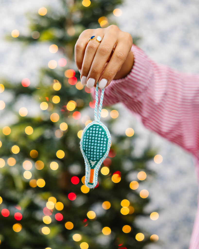Hand holding a cross-stitched ornament in front of a decorated Christmas tree.