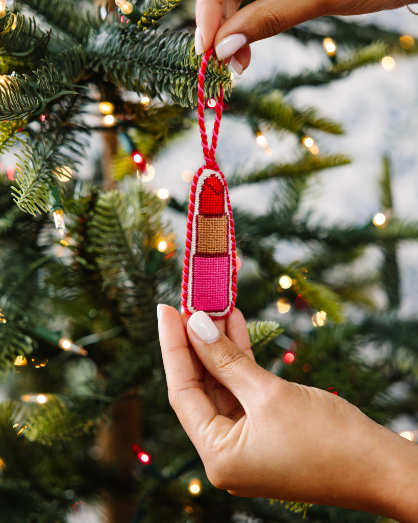 Hand holding a small ornament shaped like a lipstick against a Christmas tree background