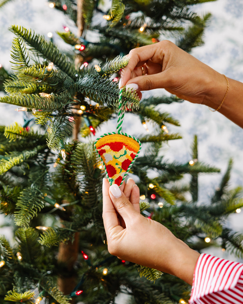 Hand holding a pizza-shaped ornament in front of a decorated Christmas tree.