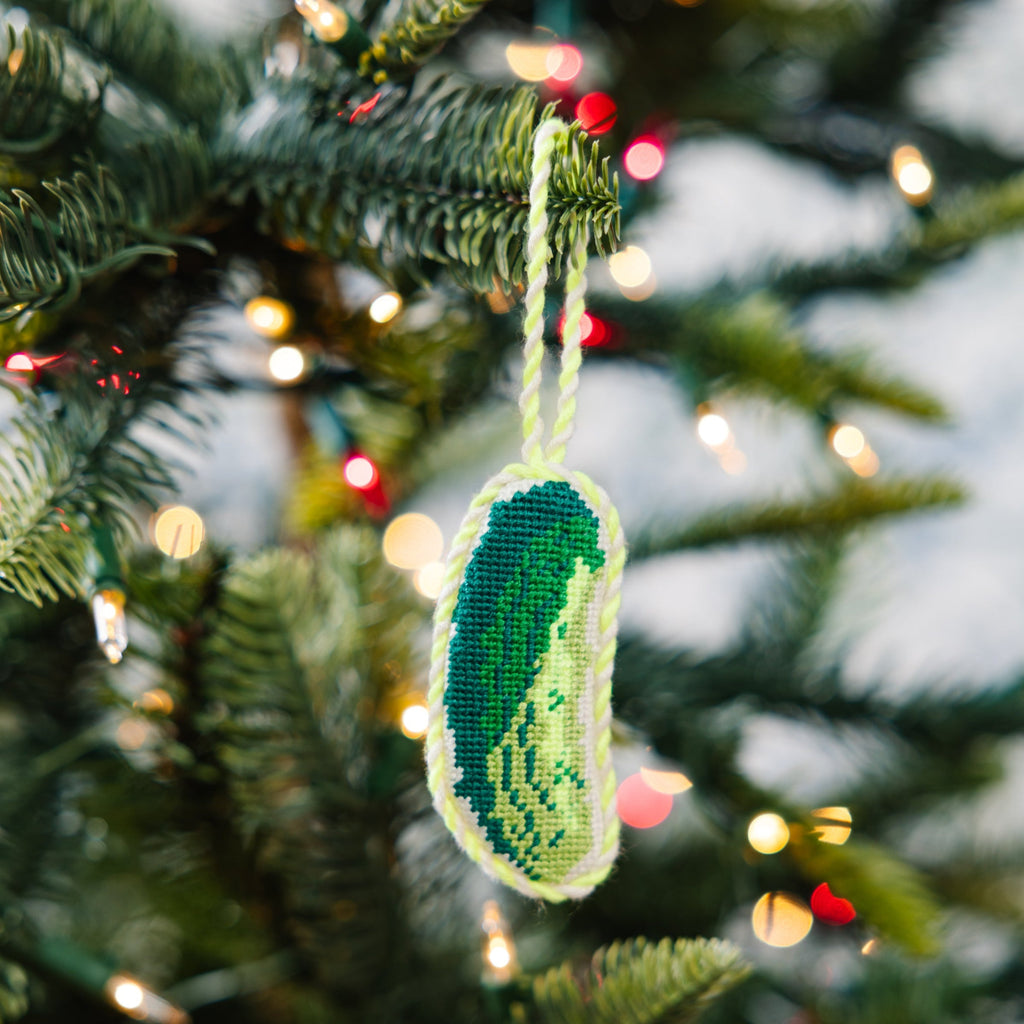 Decorative ornament shaped like a pickle hanging on a Christmas tree with lights in the background.