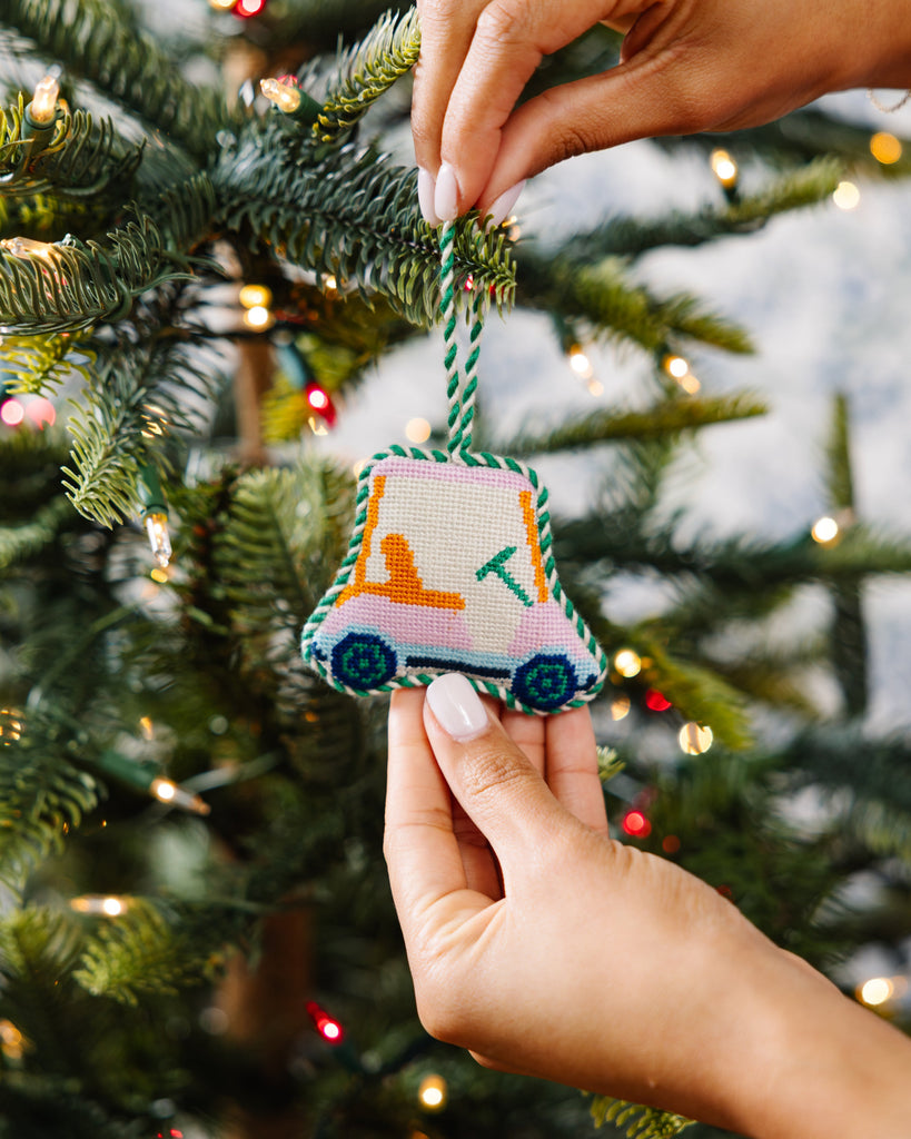 Hand holding a small embroidered ornament shaped like a car against a Christmas tree background.