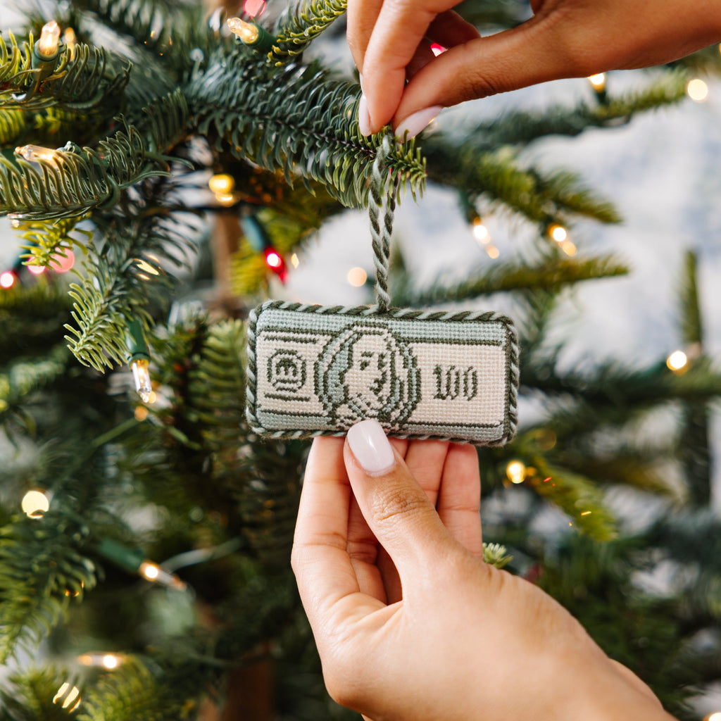 Hand holding a small decorative item shaped like a dollar bill against a Christmas tree background.