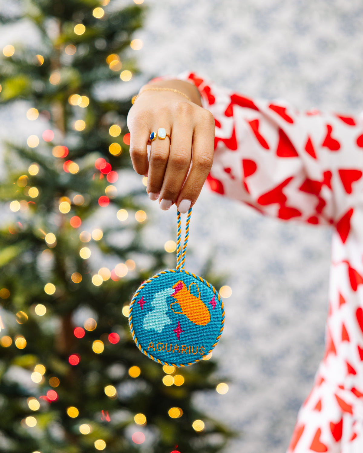Person holding a Christmas ornament with a zodiac sign design in front of a decorated tree.