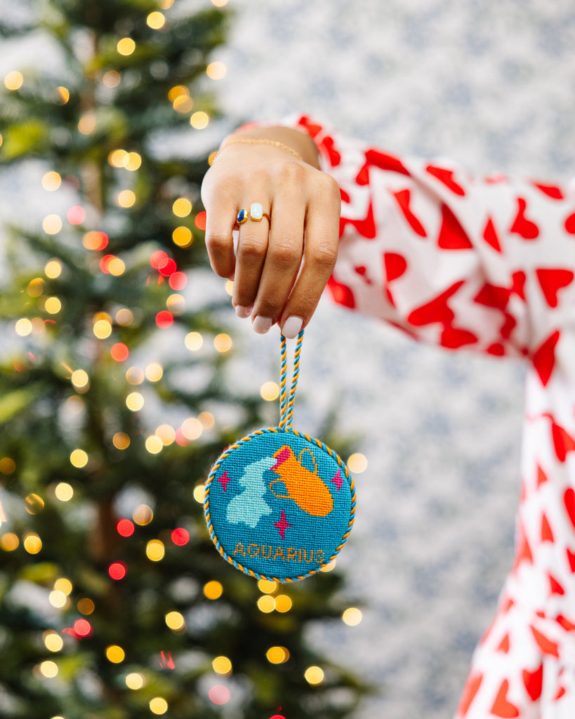Person holding a Christmas ornament with a zodiac sign design in front of a decorated tree.