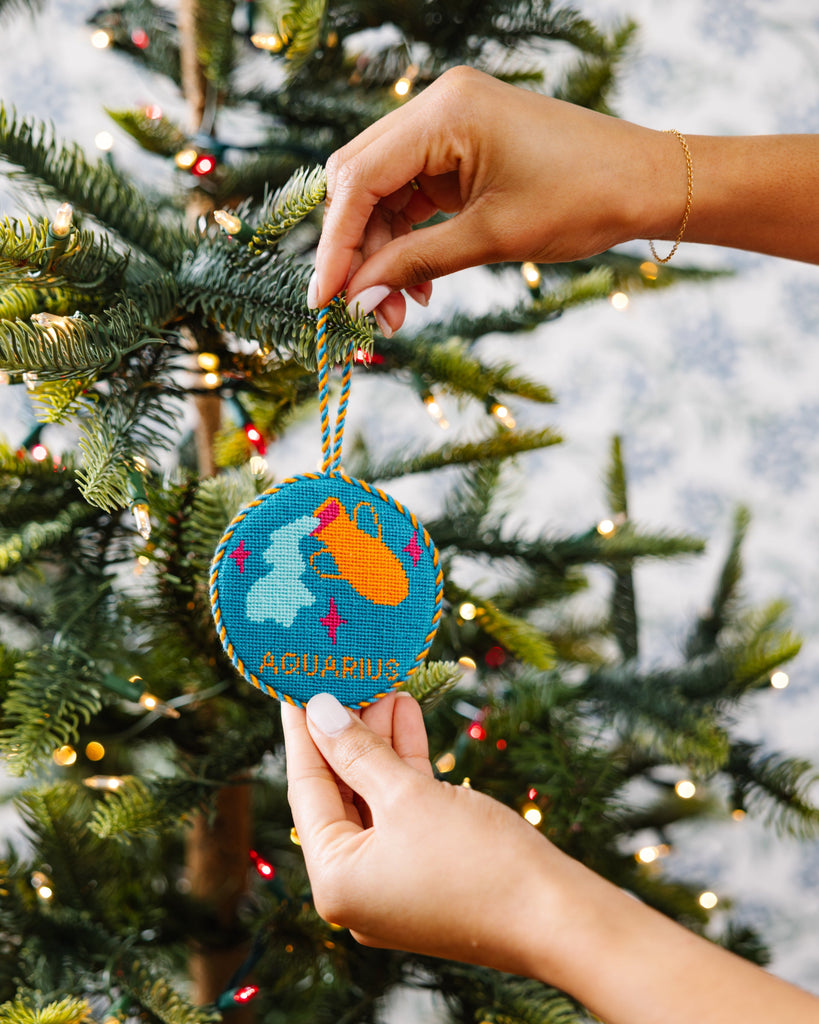 Hand holding a 'Aquarius' ornament in front of a decorated Christmas tree.