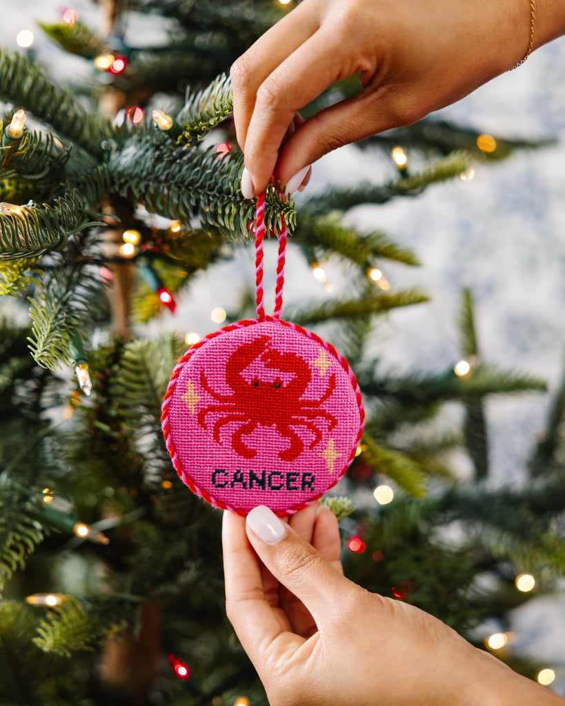 Hand holding a pink 'Cancer' zodiac ornament against a Christmas tree background