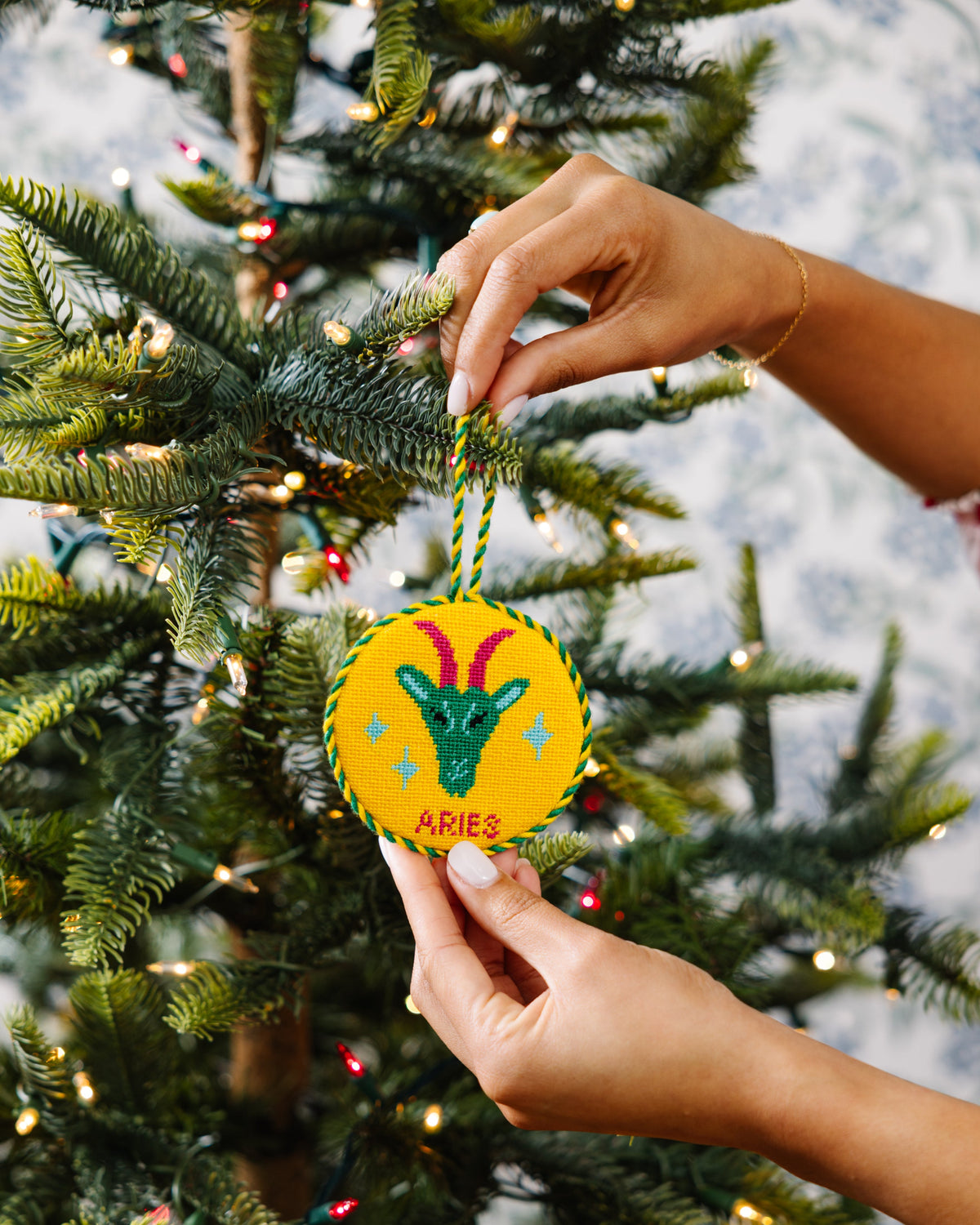 Hand holding a 'Aries' themed Christmas ornament against a decorated tree background
