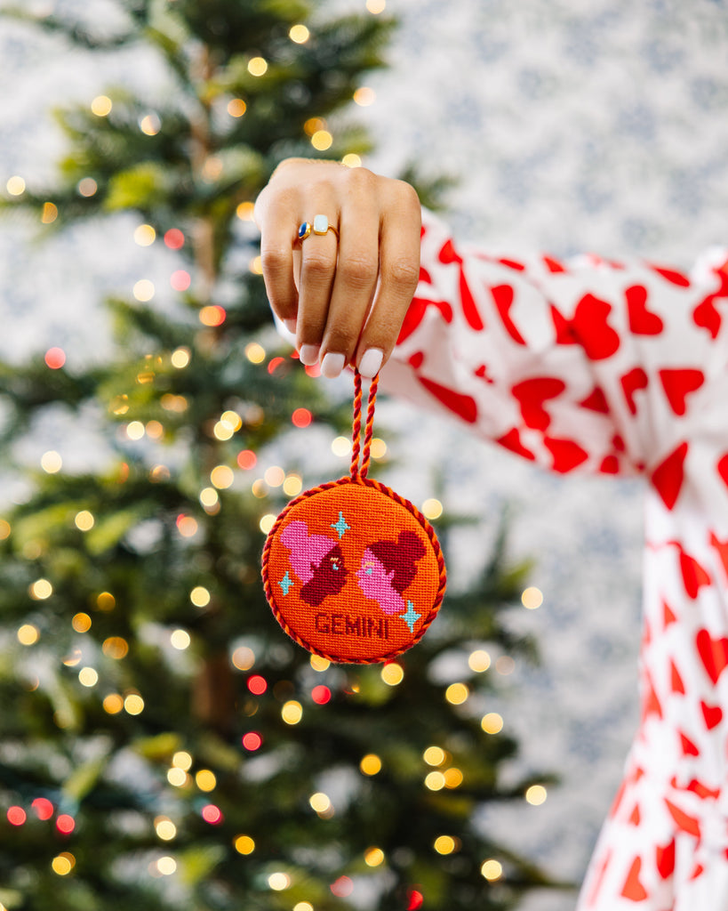 Hand holding a red ornament with 'Gemini' text in front of a decorated Christmas tree.