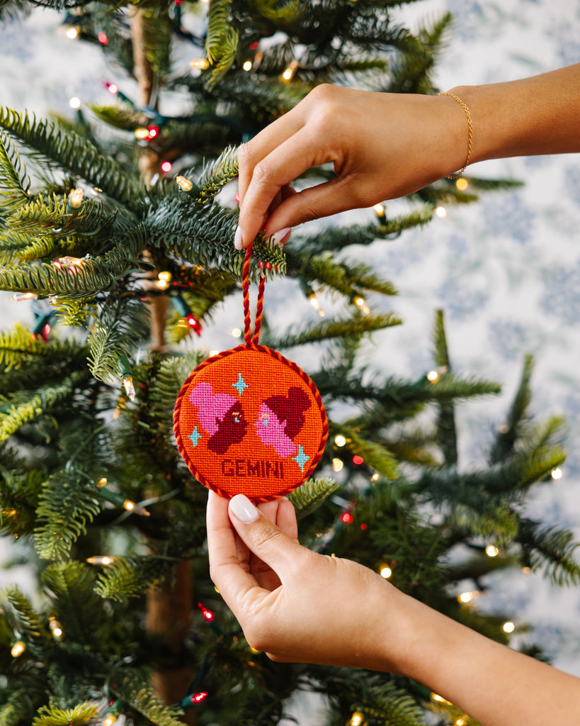 Hand holding a red embroidered ornament with 'GEMINI' on a decorated Christmas tree.