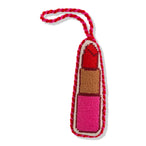 A needlepoint ornament shaped like a lipstick with a red, beige, and pink design, backed with red velvet and tied with a red ribbon.