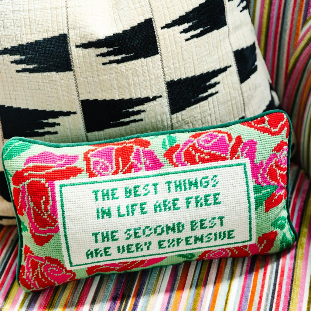 a closer look of a hand embroidered in wool and backed in luxe green velvet chic needlepoint pillow with "The best things in life are free, The second best are very expensive" cheeky saying in front sitting on a colorful couch with a black and white throw pillow at the back