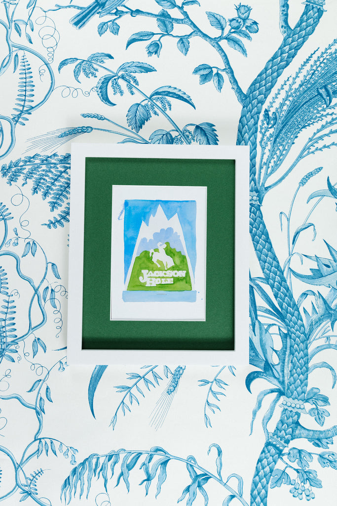 Jackson Hole Matchbook - Furbish Studio, Jackson Hole matchbook watercolor print illustrating the town's scenery painted in light blue and mud green in a 5x7 white frame with a wallpaper background