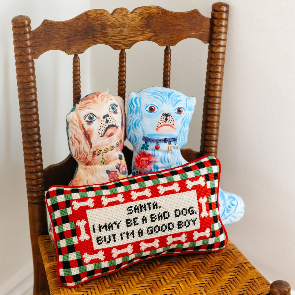 hand embroidered in wool and backed in luxe red velvet chic needlepoint pillow with "Santa, I may be a bad dog but I'm a good boy" cheeky saying in front sitting on chair with a brown and blue dog design pillows at the back of it