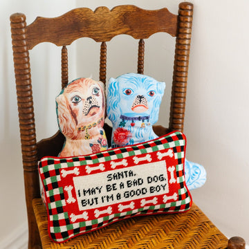 hand embroidered in wool and backed in luxe red velvet chic needlepoint pillow with "Santa, I may be a bad dog but I'm a good boy" cheeky saying in front sitting on chair with a brown and blue dog design pillows at the back of it