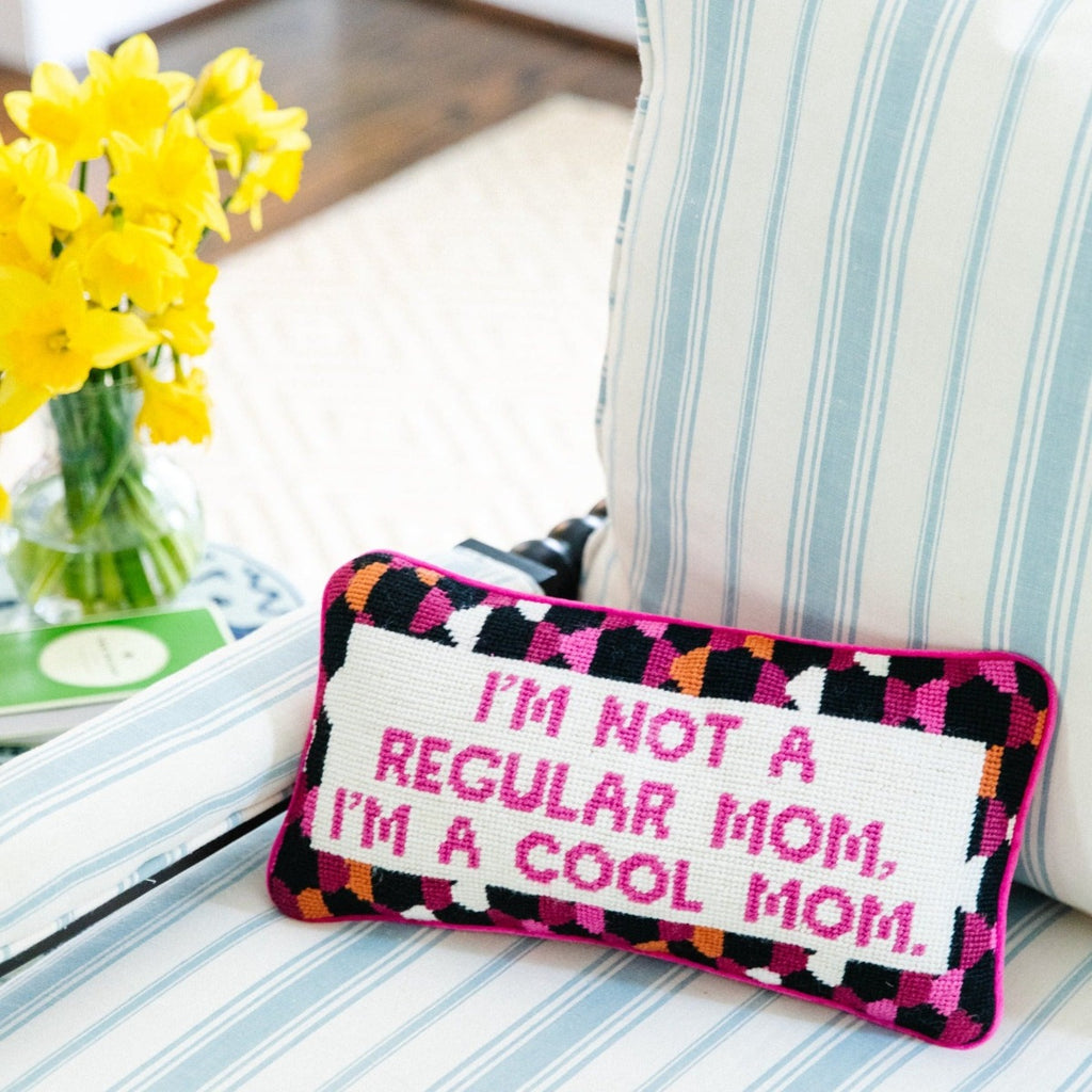 frontal view of a luxe hot pink chic hand needlepoint pillow with "I'm not a regular mom, I'm a cool mom" saying in front while sitting on a chair