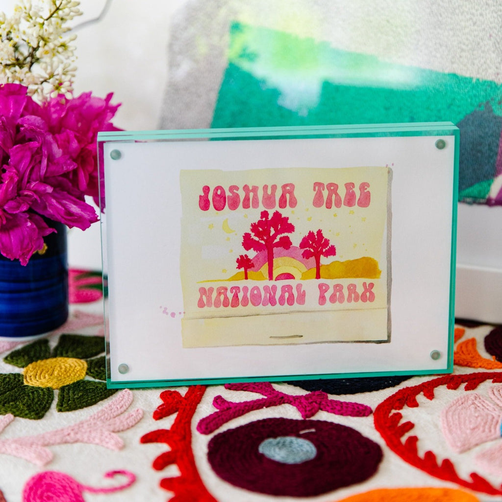Joshua Tree Matchbook - Furbish Studio, "Joshua Tree National Park" matchbook watercolor print illustrating the park's beautiful scenery painted in a pink and yellow shade enclosed in a 5" x 7" mint green magnetic acrylic floating frame on top of a table