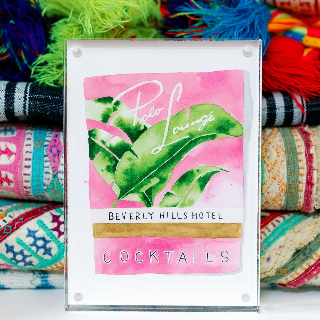 Beverly Hills Matchbook - Furbish Studio, Beverly Hills Hotel Photo Lounge matchbook watercolor print featuring a collection of Banana leaves with a pink background enclosed in a 5" x 7" see-through magnetic acrylic floating frame  and a group of folded linens behind it